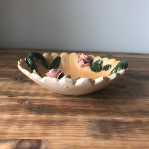 May include: A small, white ceramic bowl with a scalloped edge and a floral design. The bowl has three pink roses and green leaves.