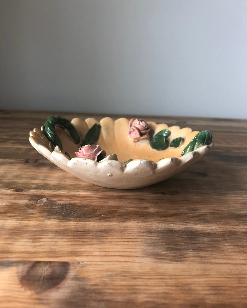 May include: A small, white ceramic bowl with a scalloped edge and a floral design. The bowl has three pink roses and green leaves.