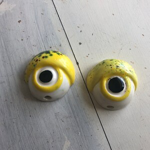 Könnte beinhalten: Zwei gelbe Keramik-Wandhaken in Form von Augen mit schwarzen Pupillen und weißen Iris. Die Augen haben grüne und braune Flecken auf der gelben Oberfläche.