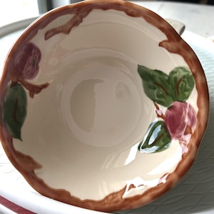 May include: Cream-colored ceramic bowl with a brown rim and floral design. The bowl features a brown border with stylized branches and red and green floral accents. The interior is a light cream color.