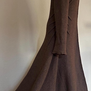 May include: A long, dark brown knit coat with a hood. The coat features long sleeves and a flared skirt. The fabric has a ribbed texture, and the coat appears to be made of a warm material, suitable for cooler weather.