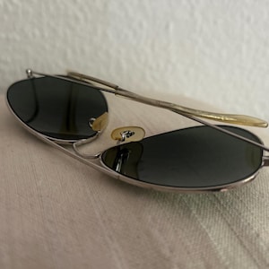 Puede incluir: Gafas de sol de aviador con montura plateada y lentes oscuras. Las gafas tienen un dise&ntilde;o cl&aacute;sico con doble puente y varillas curvas. Las lentes son de forma ovalada y la montura es de metal.