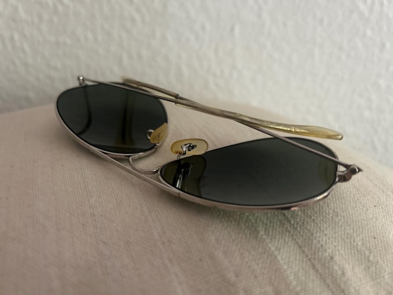 Puede incluir: Gafas de sol de aviador con montura plateada y lentes oscuras. Las gafas tienen un dise&ntilde;o cl&aacute;sico con doble puente y varillas curvas. Las lentes son de forma ovalada y la montura es de metal.