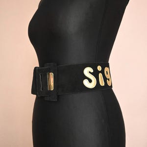 Op de afbeelding: Een zwarte riem met een gouden gesp en het woord "Sig" in gouden letters. De riem wordt getoond op een zwarte mannequin. De riem is gemaakt van een suède-achtig materiaal.