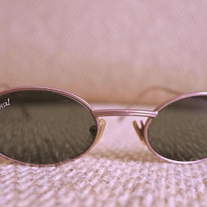 Könnte beinhalten: Ovale Sonnenbrille mit einem hellrosa Metallrahmen und dunkelgrünen Gläsern. Das Wort "Fossil" ist auf dem linken Glas aufgedruckt. Die Sonnenbrille hat ein klassisches, Vintage-Design.