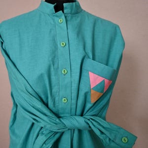 Könnte beinhalten: Ein türkisfarbenes Hemd mit Stehkragen und langen Ärmeln. Das Hemd hat eine Tasche mit einem geometrischen Design in Rosa, Orange und Türkis. Die Ärmel sind in der Taille geknotet und das Hemd ist vorne geknöpft.