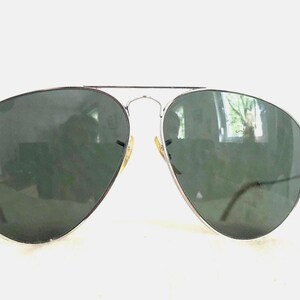 Puede incluir: Gafas de sol de aviador con montura de metal plateado y lentes verde oscuro. Las lentes tienen forma de l&aacute;grima. Las gafas de sol est&aacute;n sobre un fondo blanco.