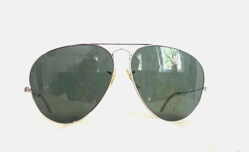 Puede incluir: Gafas de sol de aviador con montura de metal plateado y lentes verde oscuro. Las lentes tienen forma de l&aacute;grima. Las gafas de sol est&aacute;n sobre un fondo blanco.