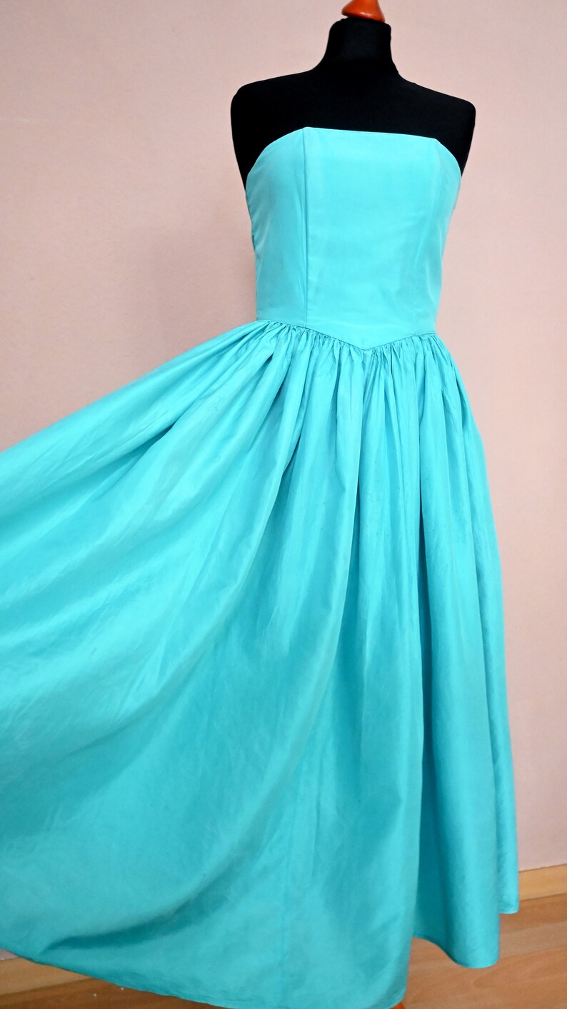 Peut inclure: Une robe turquoise sans bretelles avec un corsage ajust&eacute; et une jupe ample. La robe est faite d'un tissu lisse et soyeux.