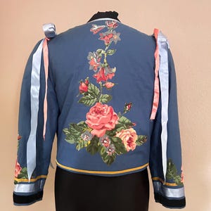 May include: A blue jacket featuring a large floral print on the back with red and pink roses and green leaves. The sleeves are adorned with light blue ribbons and a yellow trim along the hem. The jacket has a black collar and black cuffs.