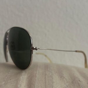 Puede incluir: Gafas de sol estilo aviador con montura plateada y lentes verde oscuro. Las gafas descansan sobre una superficie texturizada de color blanquecino. Las patillas tienen una ligera curva y una punta de color claro.