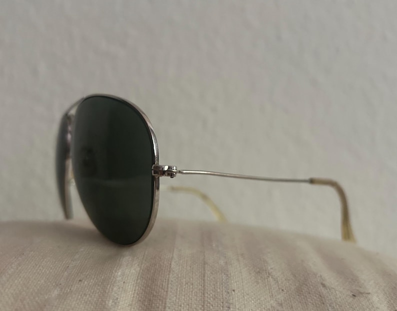 Puede incluir: Gafas de sol estilo aviador con montura plateada y lentes verde oscuro. Las gafas descansan sobre una superficie texturizada de color blanquecino. Las patillas tienen una ligera curva y una punta de color claro.