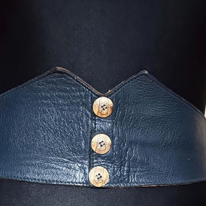 Könnte beinhalten: Ein marineblauer Ledergürtel mit einem breiten, konturierten Design. Der Gürtel hat drei goldfarbene Knöpfe in der Mitte. Der Gürtel wird auf einer schwarzen Schaufensterpuppe präsentiert.
