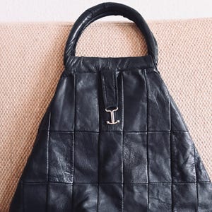 Op de afbeelding: Zwarte leren handtas met een gestructureerd, gewatteerd ontwerp. De tas heeft een ronde handgreep en een zilverkleurige sluiting met een 'I'-detail. De tas staat op een lichtroze ondergrond.