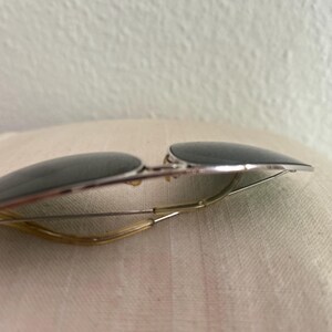 Puede incluir: Gafas de sol estilo aviador con lentes oscuras y montura de metal plateado. Las gafas tienen un dise&ntilde;o cl&aacute;sico con un puente fino y curvado y varillas. Las lentes son de color gris oscuro y la montura tiene un acabado brillante.