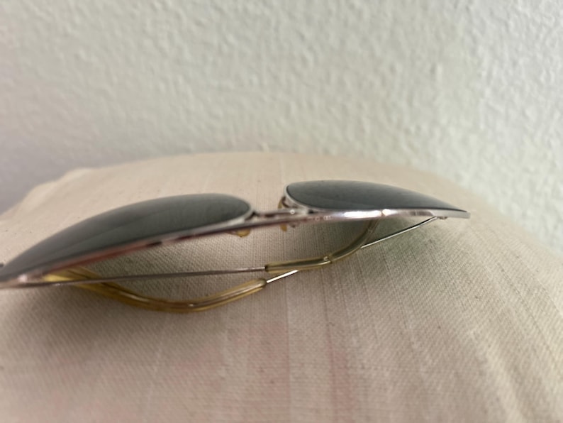 Puede incluir: Gafas de sol estilo aviador con lentes oscuras y montura de metal plateado. Las gafas tienen un dise&ntilde;o cl&aacute;sico con un puente fino y curvado y varillas. Las lentes son de color gris oscuro y la montura tiene un acabado brillante.