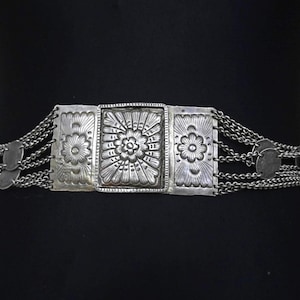 May include: Silver-coloured belt with a rectangular buckle featuring a floral design. The belt has three rectangular panels with floral patterns and is connected by chains and small, round, decorative elements. The belt is worn on a black mannequin.