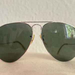 Puede incluir: Gafas de sol estilo aviador con montura de metal plateado y lentes verdes. Las lentes en forma de l&aacute;grima est&aacute;n engastadas en una montura fina plateada con doble puente. Las patillas son finas y curvadas. Un accesorio cl&aacute;sico.