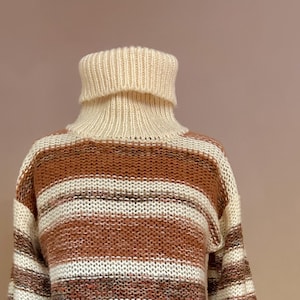 May include: A chunky knit turtleneck sweater featuring horizontal stripes in shades of cream, tan, and brown. The garment has a thick, ribbed collar and long sleeves, constructed with a loose, textured weave pattern throughout the body.