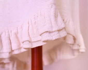 May include: Close-up of a cream-colored fabric with a textured weave. The fabric features a layered ruffle design along the bottom edge. The ruffles add a decorative element to the textile.