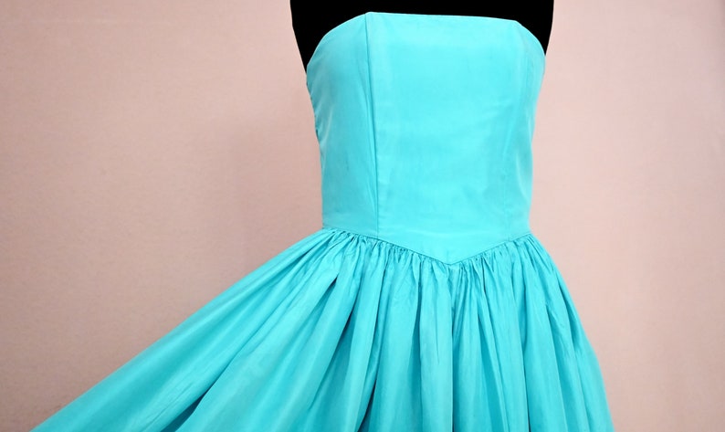 Peut inclure: Une robe turquoise sans bretelles avec un corsage ajust&eacute; et une jupe ample. La robe est faite d'un tissu lisse et soyeux.