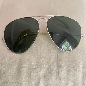 Puede incluir: Gafas de sol de aviador con montura de metal plateado y lentes verde oscuro. Se aprecia la cl&aacute;sica forma de l&aacute;grima. Las gafas de sol est&aacute;n sobre una superficie clara y texturizada.