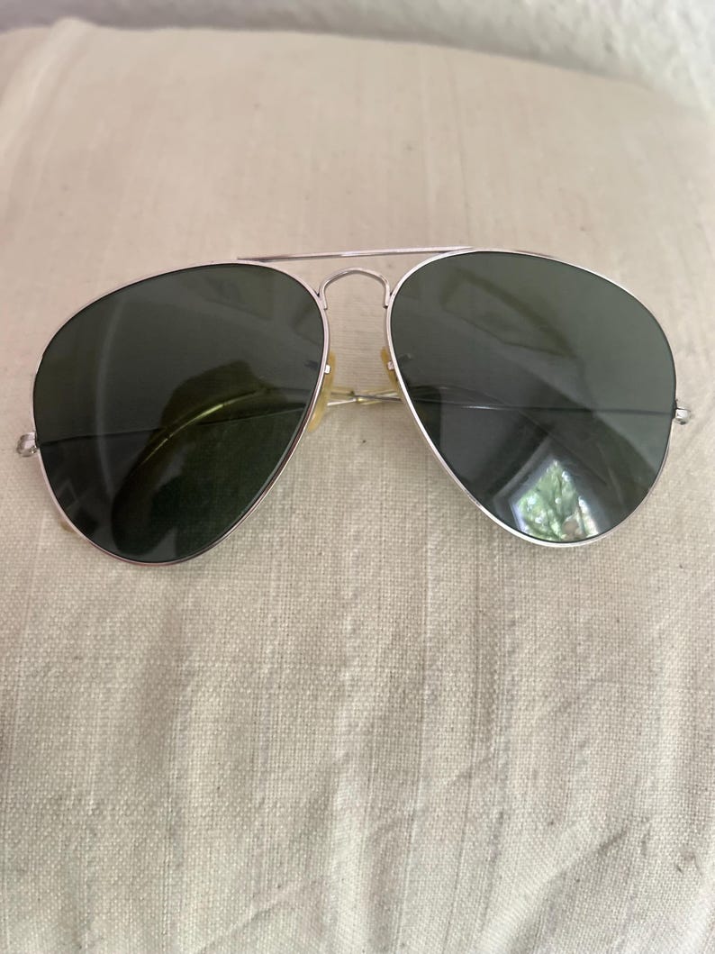 Puede incluir: Gafas de sol de aviador con montura de metal plateado y lentes verde oscuro. Se aprecia la cl&aacute;sica forma de l&aacute;grima. Las gafas de sol est&aacute;n sobre una superficie clara y texturizada.