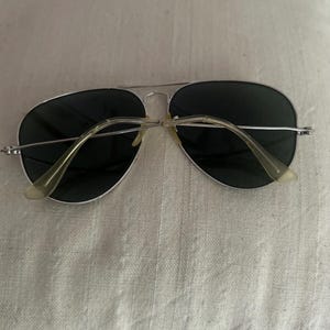 Puede incluir: Gafas de sol de aviador con montura plateada y lentes oscuras. Las gafas tienen un doble puente y patillas de color claro. Las lentes son de color verde oscuro.