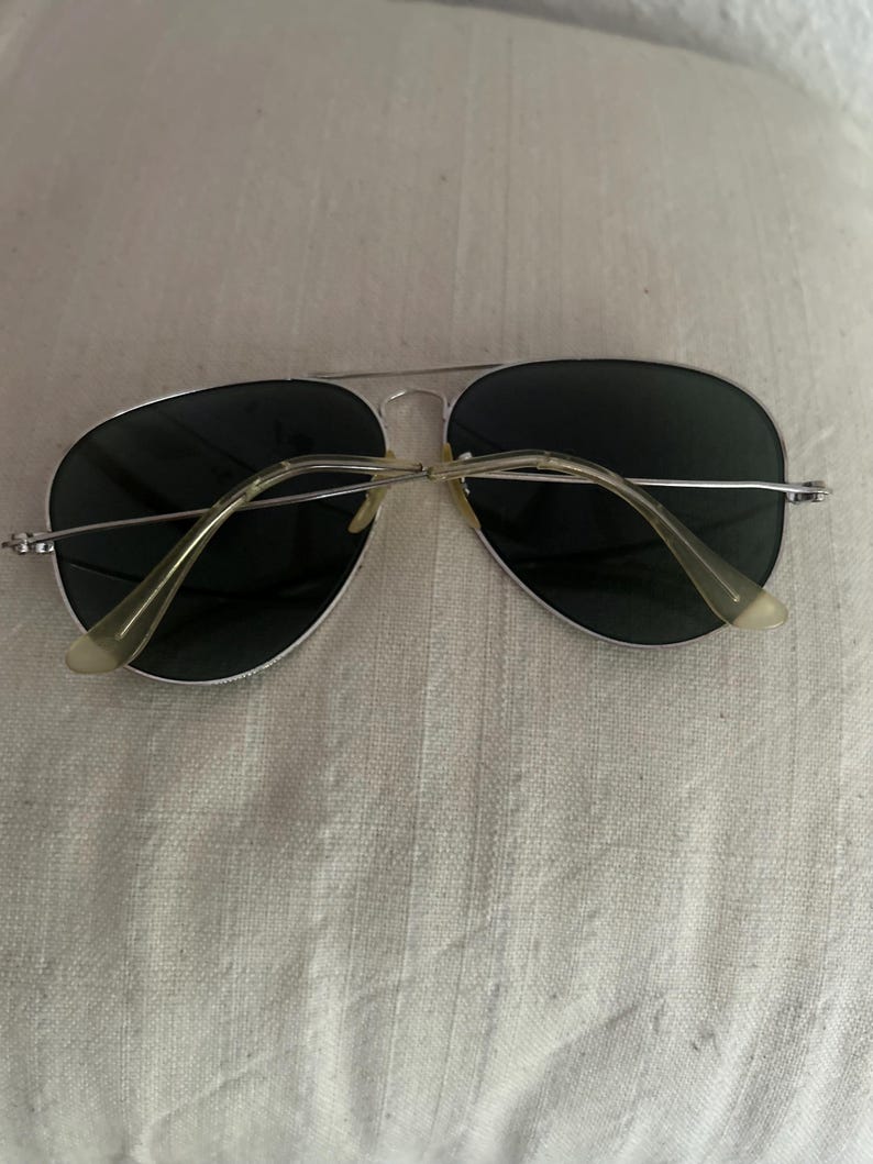 Puede incluir: Gafas de sol de aviador con montura plateada y lentes oscuras. Las gafas tienen un doble puente y patillas de color claro. Las lentes son de color verde oscuro.