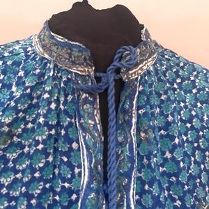 Op de afbeelding: Een blauwe blouse met een herhalend bloemenpatroon in turquoise en witte tinten. Het kledingstuk heeft een hoge kraag met een centrale verticale sluiting en een blauwe gevlochten koordsluiting bij de halslijn voor een geplooid effect.
