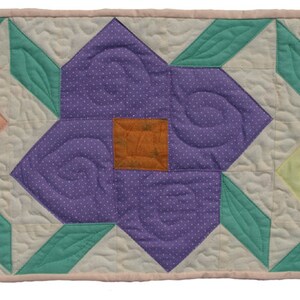 May include: A quilted table runner featuring three large, stylized flowers in shades of peach, purple, and yellow, each with an orange center. The flowers are set against a cream background with green leaves and a pink border. The quilting creates a textured effect.