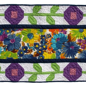 May include: A quilted table runner featuring a central floral pattern in shades of blue, yellow, and green. The runner is bordered by a navy blue frame and adorned with purple diamond-shaped flower blocks and green leaf vines on a white background.