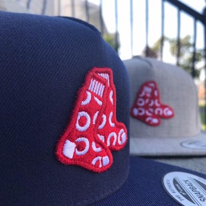 May include: A navy blue snapback cap featuring an embroidered red sock logo with white circular patterns. In the background, a light gray snapback cap displays the same red and white sock emblem. Both caps have a flat brim and a structured crown.