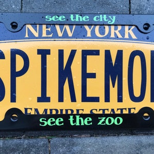 May include: A black license plate frame with a yellow background. The text "NEW YORK" is at the top of the frame. The text "SPIKEMON" is in large, bold, black letters in the center of the frame. The text "EMPIRE STATE" is at the bottom of the frame. The text "see the city" is at the top of the frame in green letters. The text "see the zoo" is at the bottom of the frame in green letters.