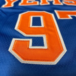 Puede incluir: Una camiseta de baloncesto azul con el texto YEMS en naranja y ribete blanco. La camiseta muestra un gran n&uacute;mero 9 naranja con detalles de costura en blanco y azul. La tela tiene una textura de malla perforada en toda la prenda.