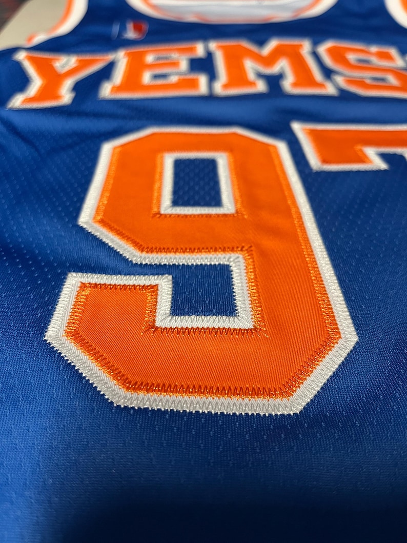 Puede incluir: Una camiseta de baloncesto azul con el texto YEMS en naranja y ribete blanco. La camiseta muestra un gran n&uacute;mero 9 naranja con detalles de costura en blanco y azul. La tela tiene una textura de malla perforada en toda la prenda.