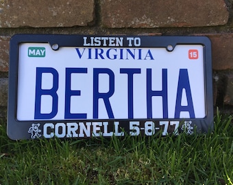 Puede incluir: Marco de matrícula negro con fondo blanco. El marco tiene el texto "Listen to Virginia" en la parte superior, "Bertha" en letras azules en el centro y "Cornell 5-8-77" con un gráfico de oso bailando en la parte inferior.