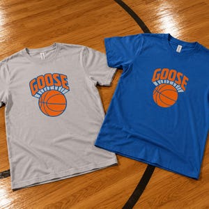 May include: A light gray t-shirt and a blue t-shirt, each featuring a graphic print on the chest. The graphic consists of the word GOOSE in orange letters above an orange basketball with black lines, all outlined in white.