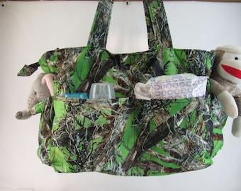 May include: A green camouflage print nappy bag with multiple pockets. The bag has a zipped top closure and two long handles. The bag is filled with various baby items, including a stuffed animal, a bottle, and nappies.