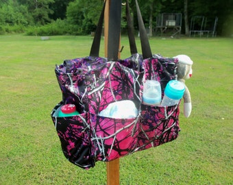 May include: A pink and black camouflage tote bag with multiple pockets. The bag is hanging from a hook and has a few items inside, including a bottle of baby formula, a bottle of baby lotion, and a stuffed monkey.