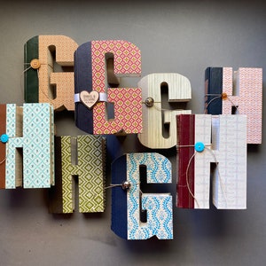 May include: A collection of decorative wooden letters, spelling out "HIGH", each adorned with patterned paper and small books. The letters are in various colors and patterns, with buttons and twine adding detail. A heart-shaped tag is attached to one letter.