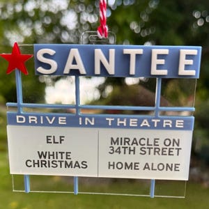 May include: A clear acrylic ornament shaped like a drive-in theater sign with a red star, blue accents, and white text reading SANTEE DRIVE IN THEATRE, listing the movies ELF, WHITE CHRISTMAS, MIRACLE ON 34TH STREET, and HOME ALONE.