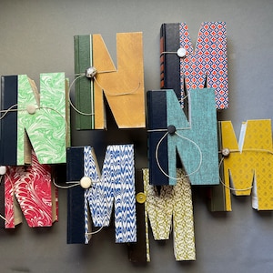 May include: An artistic arrangement of the letters "M" and "N" crafted from repurposed books. Each letter features a unique cover design, including marbled patterns, solid colors, and patterned prints. The letters are connected with twine and decorative buttons.