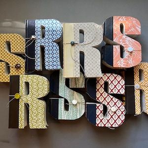 May include: A decorative wall art piece spelling out the word "SRSRSR" with large, three-dimensional letters. Each letter is crafted from repurposed books, featuring patterned covers in various colors and designs, and secured with buttons and string.