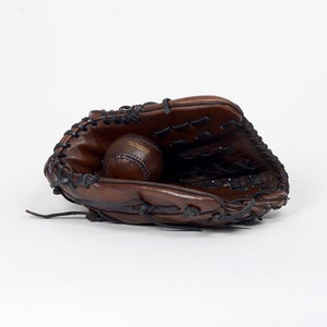 Peut inclure: Un gant de baseball en cuir marron avec une balle de baseball à l'intérieur. Le gant est ouvert et la balle repose dans la poche.