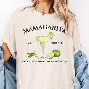 May include: A beige t-shirt with the text "Mamagarita" in black. The shirt also features a drawing of a margarita glass with limes and the text "24/7" and "Mon-Sun". The text "A cool mom who loves margaritas" is also printed on the shirt.