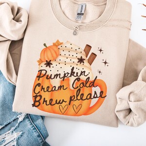 May include: A beige crewneck sweatshirt featuring a graphic of a pumpkin-shaped mug filled with whipped cream, a small pumpkin, and cinnamon sticks. The text reads Pumpkin Cream Cold Brew please, accompanied by two small heart outlines and star icons.