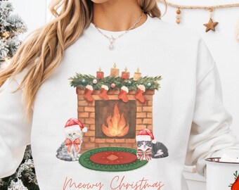 Peut inclure: Sweat-shirt blanc avec un motif de Noël. Il présente une cheminée en briques avec des bougies, des chaussettes et un feu, ainsi que deux chatons portant des chapeaux de Père Noël. L'inscription "Meowy Christmas" est écrite en cursive en dessous.