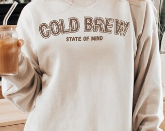 May include: A beige sweatshirt with the text "COLD BREW STATE OF MIND" printed in brown. The sweatshirt is a neutral colour and has a relaxed fit. The image shows a person holding a glass of iced coffee.