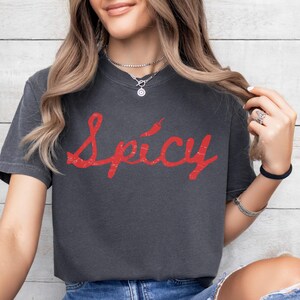 Op de afbeelding: Een donkergrijs T-shirt met een rode vintage-stijl grafische afbeelding met de tekst "Spicy" met een rode peper boven het woord.